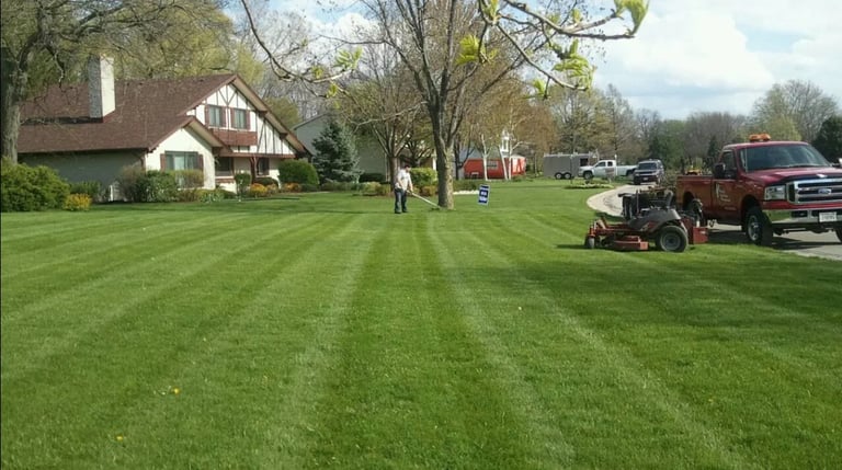 Person operating a riding lawn mower on a large striped lawn in front of a house with vehicles parked in the driveway
