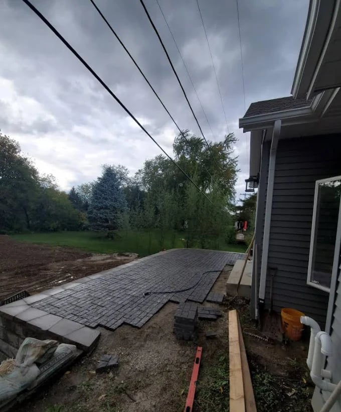 Residential patio under construction with newly laid pavers, house exterior visible on right, power lines overhead, and wooded landscape in background