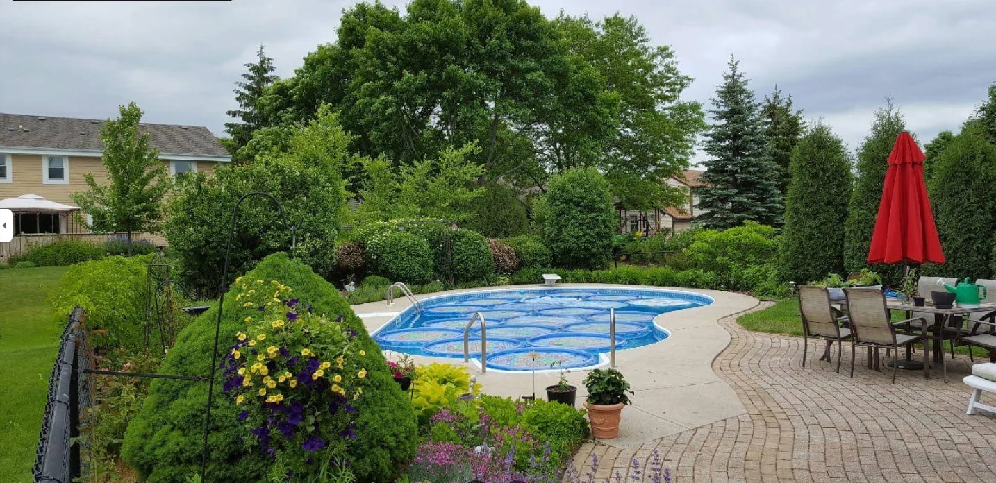 Backyard oasis with covered blue in-ground pool, brick patio, flowering gardens, lounge chairs, red umbrella, and manicured lawn surrounded by trees