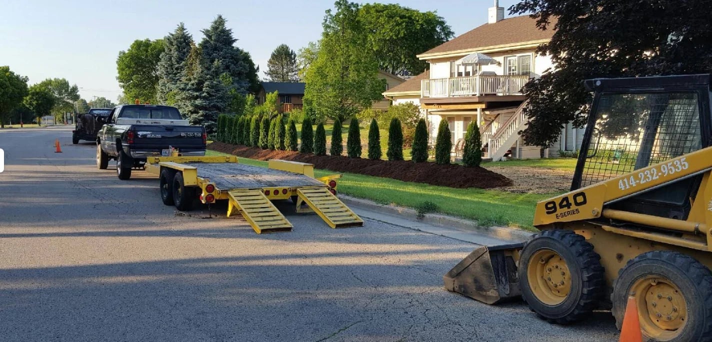 Yellow skid steer loader towing a black ramp trailer on a residential street with vehicles and houses